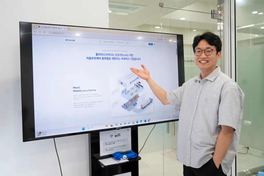 폴라리스쓰리디 이학준 이사가 대형 모니터 앞에 서서 손으로 폴라리스쓰리디 홈페이지 화면을 가리키며 설명하고 있는 모습. 화면에는 AI 로봇 기술과 관제시스템 솔루션에 대한 내용이 보인다.
